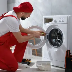Fachowiec w czerwonym ogrodniczkach pokazuje, jak podłączyć pralkę, używając śrubokręta do mocowania drzwi. Obok stoi torba z narzędziami.
