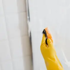 Ręka w żółtej rękawiczce czyści kabinę prysznicową gąbką. Dowiedz się, czym myć kabinę prysznicową, by lśniła czystością.