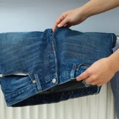 Ręce układają jeansy na grzejniku, zastanawiając się, czy jeansy można suszyć w suszarce. Obok wisi szara bluza i niebieska koszula w kratę.