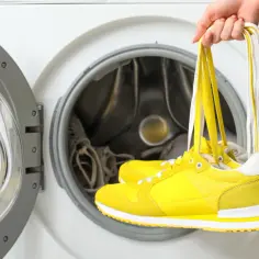 Ręka trzyma żółte buty na sznurówkach przed otwartą pralką. Pranie butów w pralce to szybki sposób na odświeżenie ulubionych sneakersów.