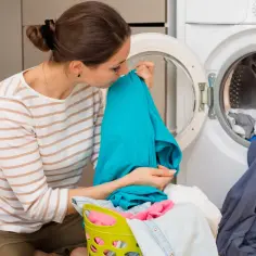 Kobieta wącha świeżo wyprane ubranie, przygotowując się do prania ręcznego w pralce.