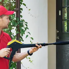 Mężczyzna w czerwonej czapce i koszulce myje elewację domowym sposobem za pomocą myjki ciśnieniowej.