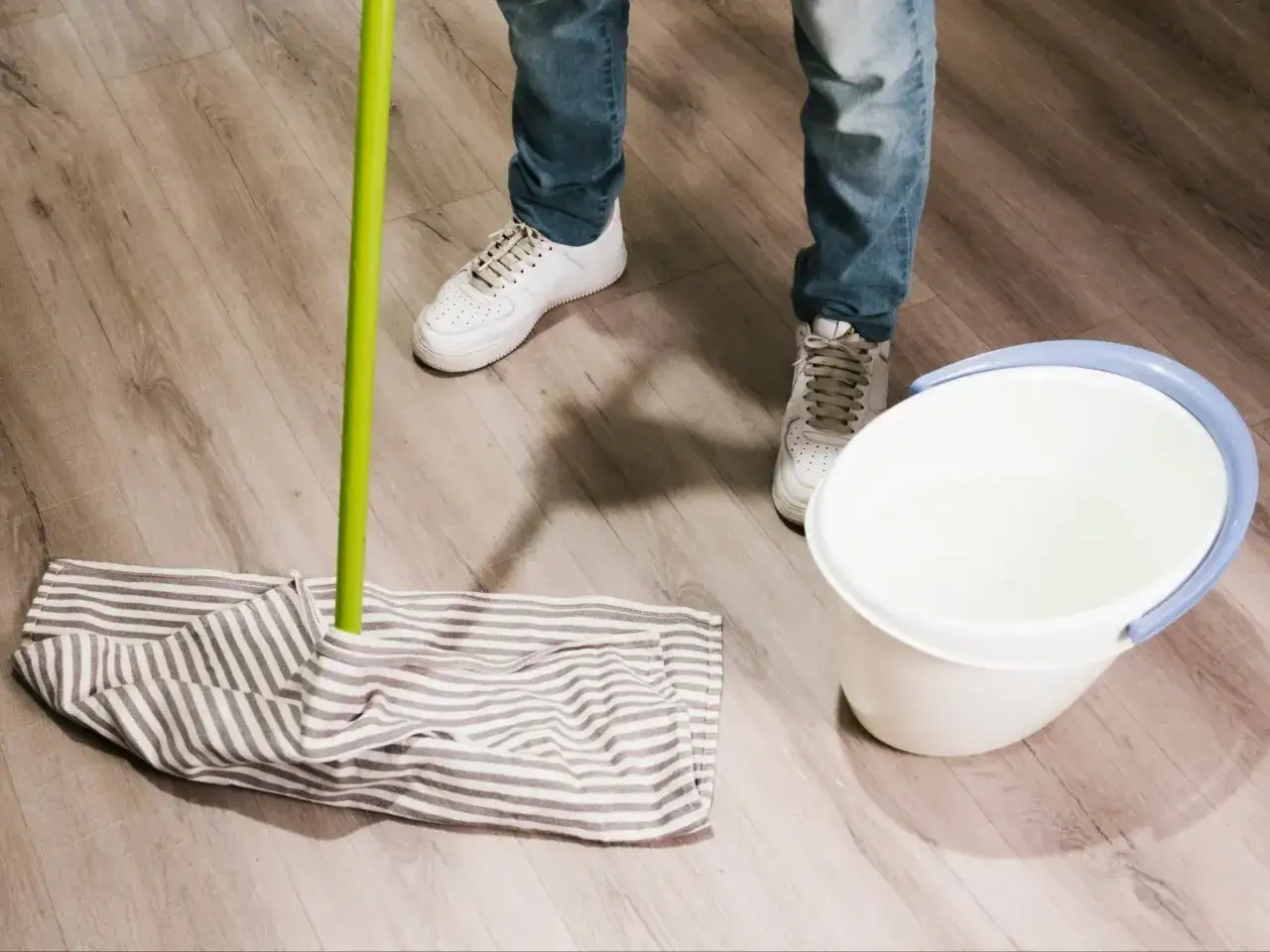 Osoba w jeansach i białych sneakersach przygotowuje się do mycia paneli. Obok mopa stoi wiadro z wodą.